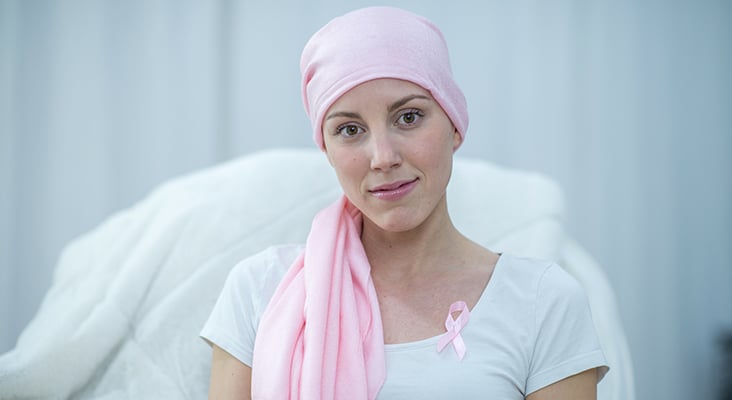 A Caucasian woman is indoors in her house. She has cancer, and she is wearing a head scarf to hide her hair loss. She is sitting in a chair and smiling at the camera.