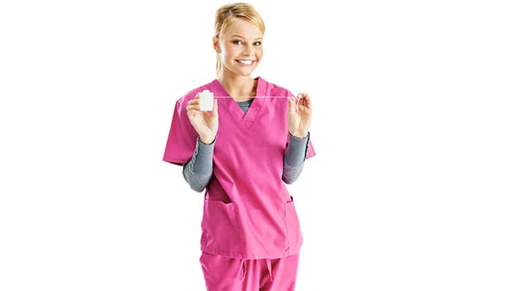 Young blonde female dental hygienist in pink scrubs, holding a piece of dental floss with a bright smile on her face; isolated on white.