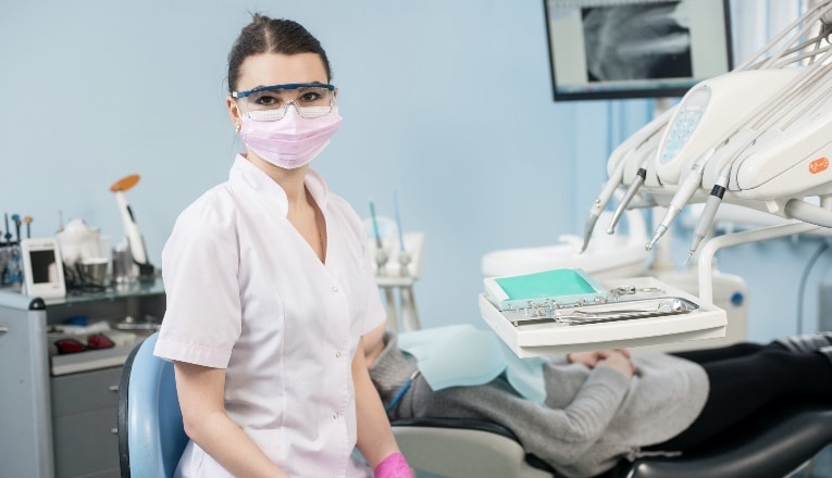 Portrait of young female dentist with patient in the dental office. Doctor wearing glasses, mask, white uniform and pink gloves.