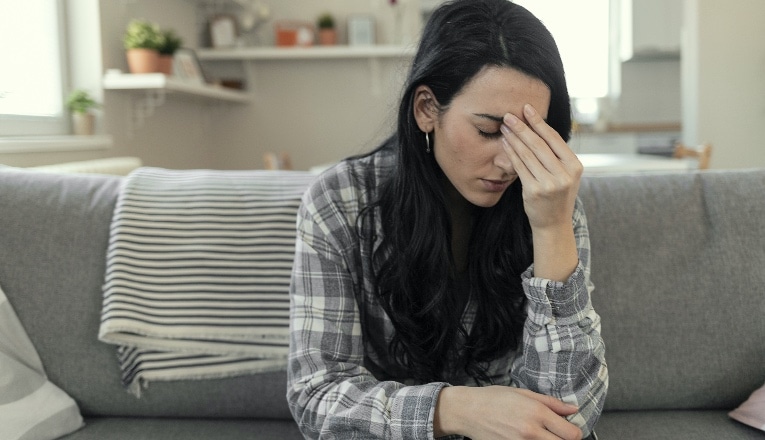 Frustrated young woman suffering from the headache while sitting on the sofa at home with an expression of being unwell, with eyes closed.