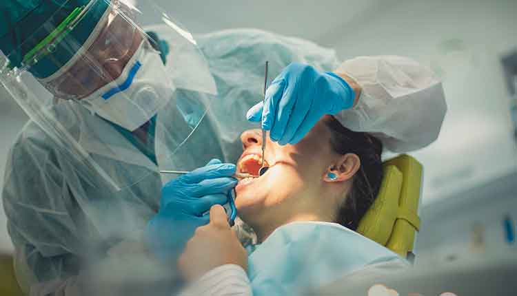 The dentist, fully dressed in a protective suit with an eyewear protection is with a patient. A woman in the chair has her mouth opened and examined while holding a suction pump.