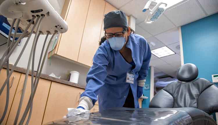 A Latina Healthcare Worker in Her Fifties Wearing a Face Mask and Surgical Gloves Wipes a Medical Chair in an Examination Room at a Dental Office in Preparation for the Next Patient