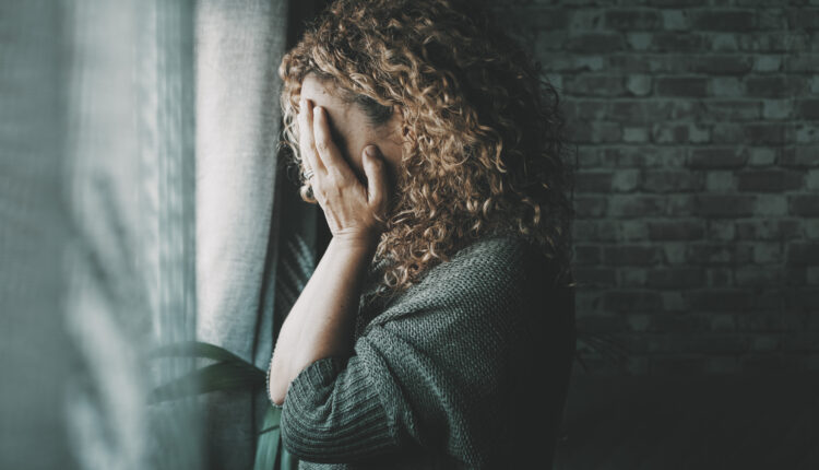Sad and worried woman side portrait at home in the dark looking outside the window. Worried and lonely female alone indoor. Concept of end of relationship and unsuccessful emotion. Lady touching face