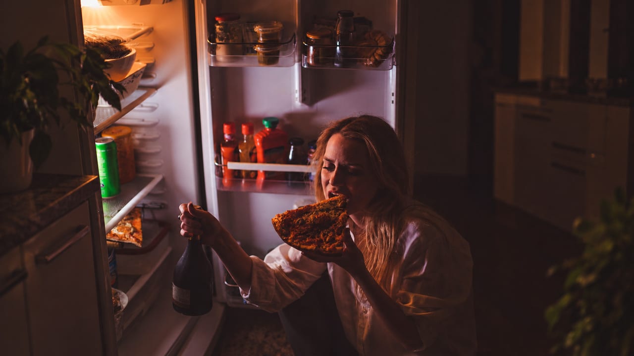 Woman eating pizza beside an open refrigerator at night